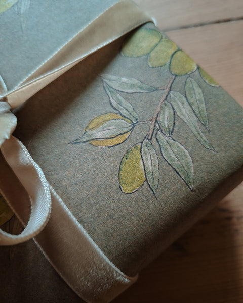 A close-up of a gift box wrapped in textured taupe paper printed with a botanical olive branch design, tied with a cream satin ribbon on a wooden surface.