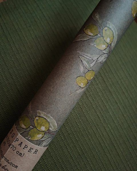 A close-up of a rolled sheet of textured kraft paper with an olive branch illustration, resting on a dark green ribbed fabric background.