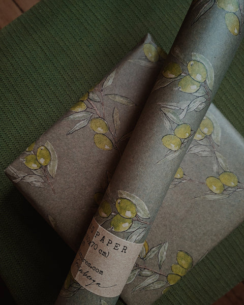 A roll of brown kraft paper wrapping paper with a watercolor print of green olives on branches, shown wrapped around gift boxes on a textured green fabric background.