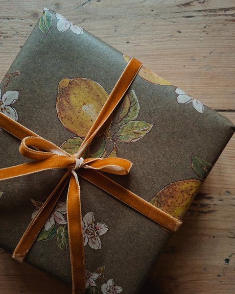 A gift box wrapped in brown kraft paper with a lemon and white flower print, tied with a wide orange velvet ribbon in a bow, set on a rustic wooden surface.