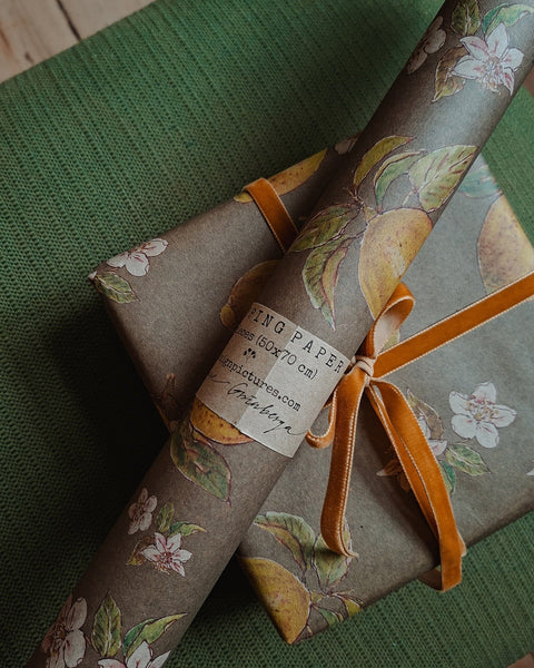 A close-up of gift wrapping paper with a vintage botanical print featuring peach, melon, and white flowers on a taupe background, wrapped around a gift box tied with a rust-orange ribbon. A paper label reads 'RING PAPER 12 X 12 X 12 cm' and 'www.