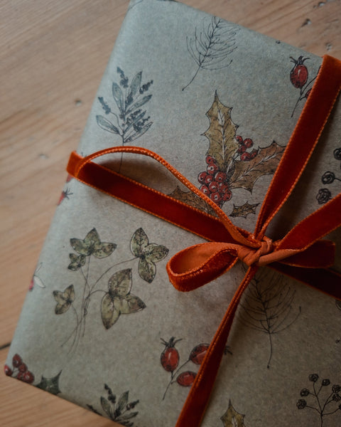 A gift box wrapped in beige kraft paper with a botanical pattern of leaves and red berries, tied with a rust-orange velvet ribbon bow, set on a light wooden surface.